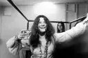 Janis Joplin z kieliszkiem w dłoni, uśmiechnięta. Jej charakterystyczne włosy i naszyjniki przywodzą na myśl jej niezapomniane utwory.