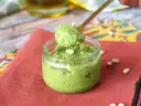 Łyżka nabiera aromatyczne pesto z rukoli z małego słoiczka. Na czerwonym tle widać orzeszki piniowe.