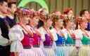 Szła dzieweczka w tradycyjnym stroju, z barwnym wiankiem na głowie, obok innych w ludowych strojach.