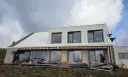 Trwa **wymiana blachy na dachu** nowoczesnego domu. Pracownicy montują nowe pokrycie, a budynek ma już zamontowane okna.