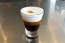 Kawa caffe coretto w szklance, z warstwami ciemnego espresso i białej pianki, ozdobiona złotą kropką.