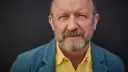 Artur Barciś w żółtej koszuli i niebieskiej marynarce, z siwą brodą, patrzy w obiektyw.