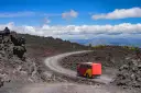 Czerwono-żółty autobus turystyczny jedzie krętą drogą przez wulkaniczne krajobrazy Etny.