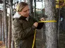 Kobieta w zielonej kurtce mierzy obwód pnia drzewa żółtą taśmą mierniczą w lesie.