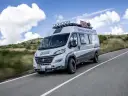 Szary kamper Ducato 4x4 Expedition z bagażnikiem dachowym i dodatkowym oświetleniem jedzie po drodze wśród wzgórz.