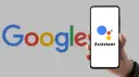 Dłoń trzyma smartfon z logo Asystenta Google na ekranie, w tle logo Google.