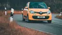 Une Twingo orange roule sur une route sinueuse bordée d'herbes sèches.