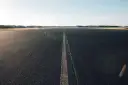 Asfaltowa droga z linią pośrodku, otoczona polami i drzewami.