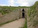 Kamienna ściana prowadzi do wejścia do grobowca Agamemnona. W oddali widać ludzi wchodzących do środka.