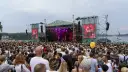 Tłum ludzi na koncercie pod gołym niebem. Na scenie gra zespół, a na ekranach wyświetlany jest green festival line up.