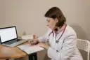 Lekarka w białym kitlu z czerwonym stetoskopem siedzi przy biurku, pisząc w zeszycie. Obok leży laptop. Czy sor wystawia L4?