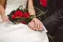 Dłonie nowożeńców splecione na tle bukietu czerwonych róż. Idealny moment, by pomyśleć, jak przygotować życzenia ślubne które zapadną w pamięć i wzruszą.