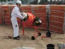Mężczyzna w białym kombinezonie wsypuje składniki do betoniarki, przygotowując beton B30. W tle palety pustaków.