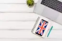 Laptop, notes, telefon z flagą Wielkiej Brytanii i długopis na białym drewnianym tle.