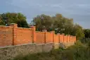 Solidny płot murowany z cegły i kamienia, z ozdobnymi słupkami, otacza teren porośnięty zielenią.