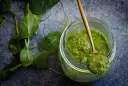 Słoik z zielonym pesto, na łyżce porcja sosu. Czy pesto ze słoika się podgrzewa? Świeże liście bazylii obok.