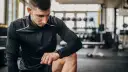 Mężczyzna w czarnej bluzie patrzy na zegarek, zastanawiając się, ile powinien trwać trening. W tle siłownia.