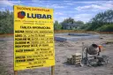 Żółta tablica informacyjna budowy z danymi o inwestycji i logo firmy LUBAR na tle placu budowy.