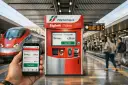 Ręka trzyma smartfon z aplikacją Trenitalia, obok biletomat na peronie. Szybkie pociągi we Włoszech czekają na pasażerów.