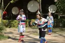 Trzy dziewczyny w tradycyjnych strojach przygotowują się do występu. W tle widać bębny, sugerujące ludową muzykę.