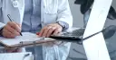 Lekarz w białym kitlu pisze na dokumentach, przygotowując zwolnienie lekarskie ze szpitala. Obok leży laptop.