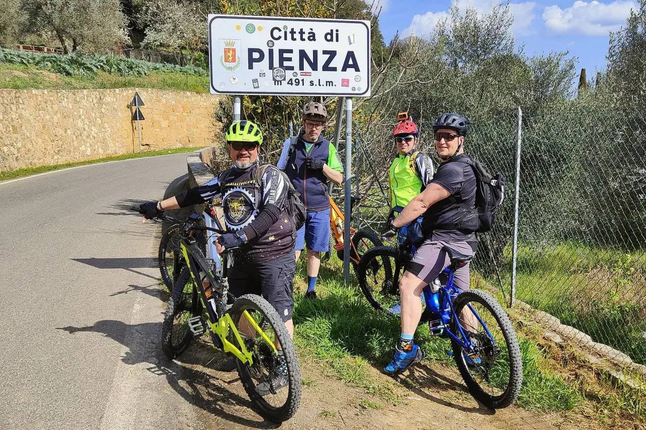 Quattro ciclisti si fermano per una foto con il cartello "Città di Pienza" durante un'avventura in bici nel Val d'Orcia.