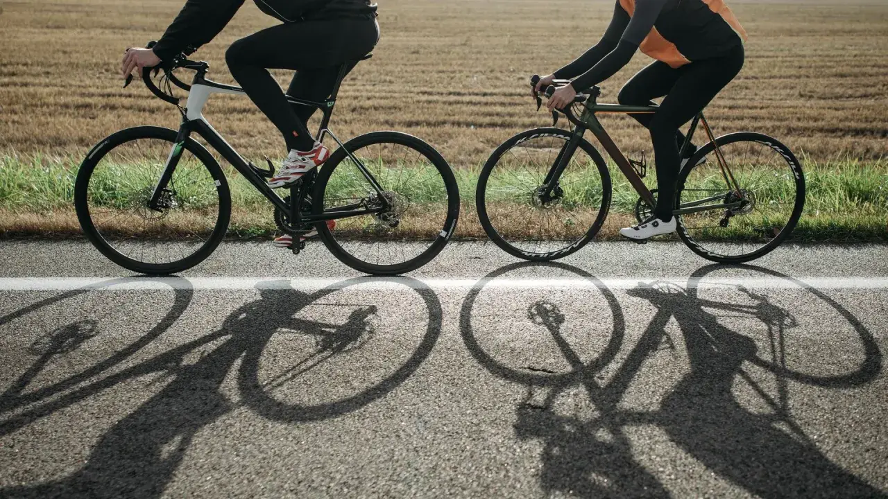 Dwie osoby na rowerach, w kaskach, jadące po drodze. Długie cienie rowerów na asfalcie.