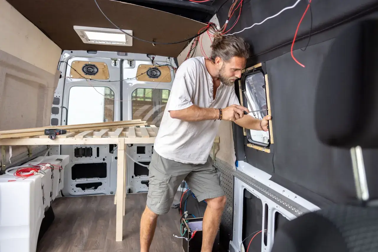 Mężczyzna buduje kamper z osobówki, montując okno. Widać drewniane elementy łóżka i instalację elektryczną.