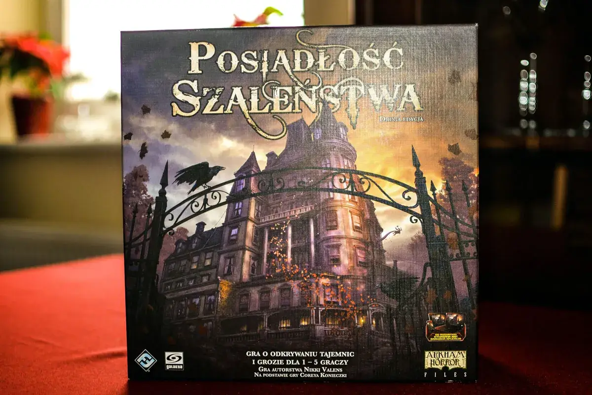 Pudełko gry "Posiadłość Szaleństwa" z mroczną posiadłością i wroną na bramie. Dodatki do tej gry planszowej zapowiadają nowe, przerażające przygody.