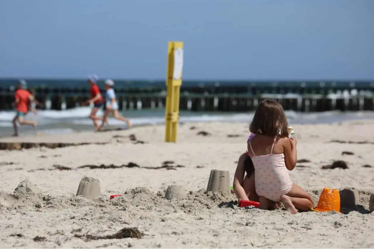 Dzieci budują zamki z piasku na plaży, w tle widać morze i falochrony. Dźwirzyno molo w oddali.