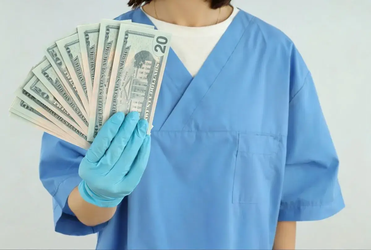 Higienistka stomatologiczna w niebieskim fartuchu pokazuje wachlarz banknotów 20 dolarowych. Zarobki netto mogą być satysfakcjonujące.