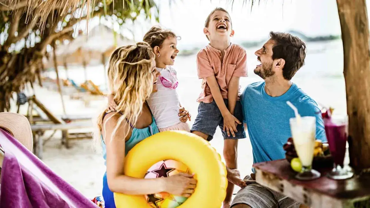 Familie lacht am Strand, genießt die Sommerferien. Ein gelber Schwimmring und Cocktails deuten auf Urlaubsfreude in den Bundesländern hin.