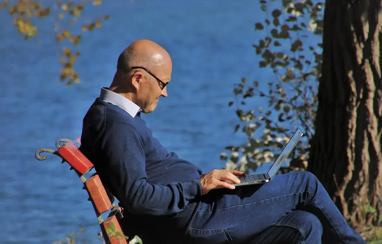 Mężczyzna w okularach siedzi na ławce nad jeziorem, pracując na laptopie. Planuje swoje ubezpieczenie emerytalne, ciesząc się spokojem natury.