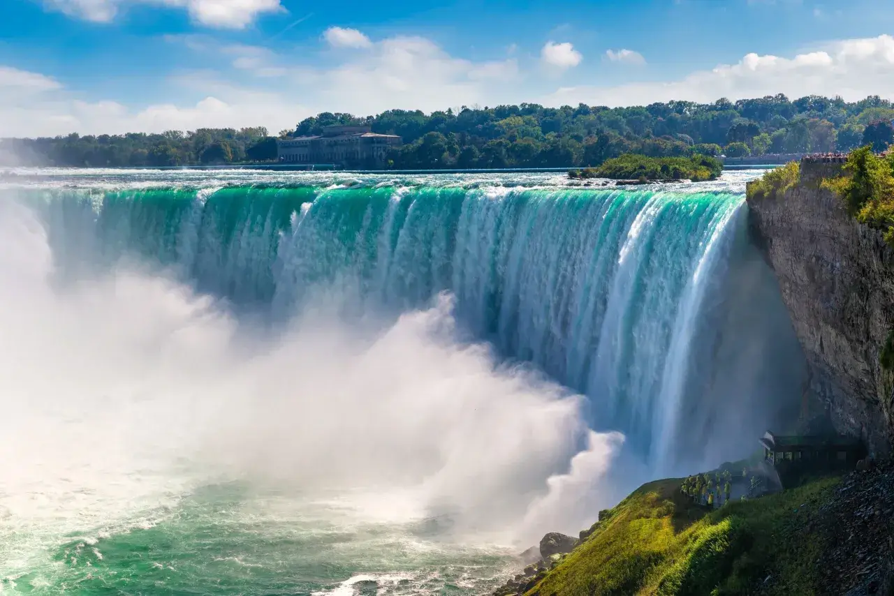 Potężny wodospad Niagara w całej okazałości. Woda spada kaskadami, tworząc mgłę. W tle drzewa i budynek.