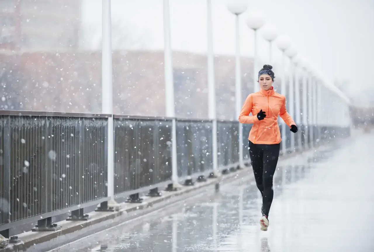 Donna corre sotto la pioggia su un ponte, con fiocchi di neve che cadono. Indossa abbigliamento sportivo arancione e nero.