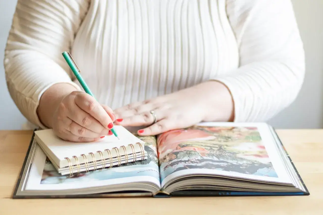 Frau schreibt mit grünem Stift in Notizbuch, während sie ein aufgeschlagenes Fotoalbum betrachtet. Sie erstellt ihr persönliches Jahrbuch.