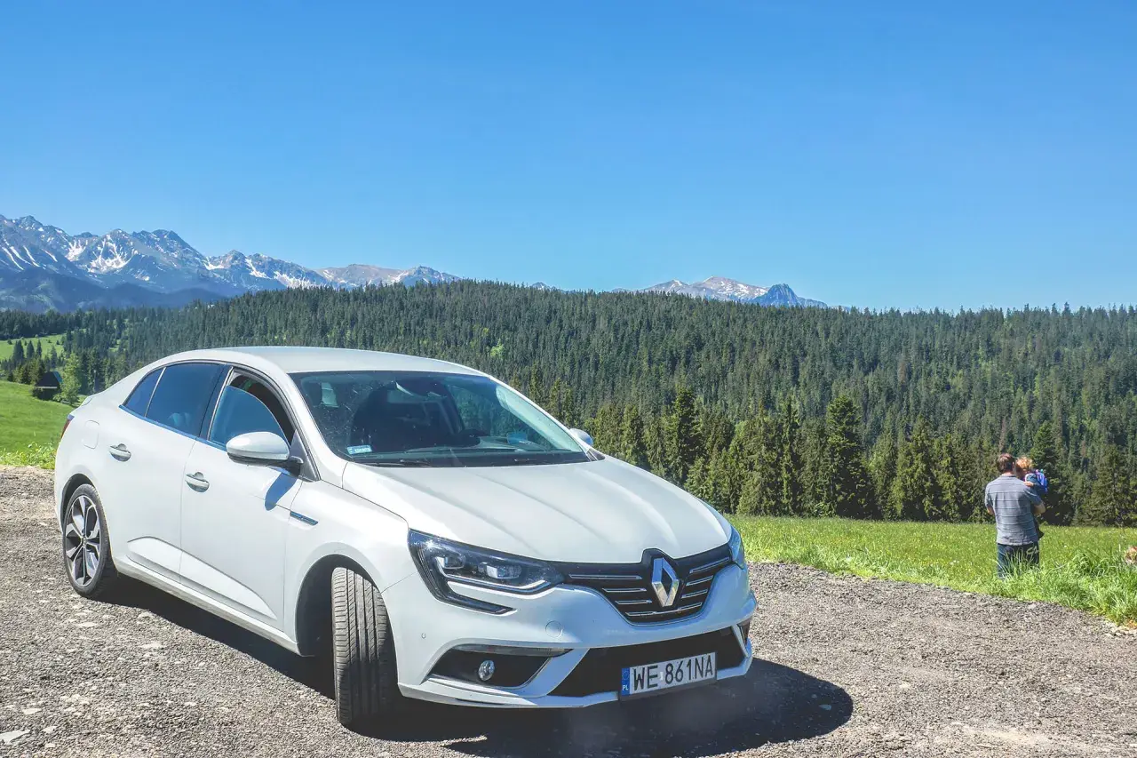 Biały Renault Megane Sedan zaparkowany na szutrowej drodze z widokiem na góry i las. Na trawie stoi mężczyzna z dzieckiem.