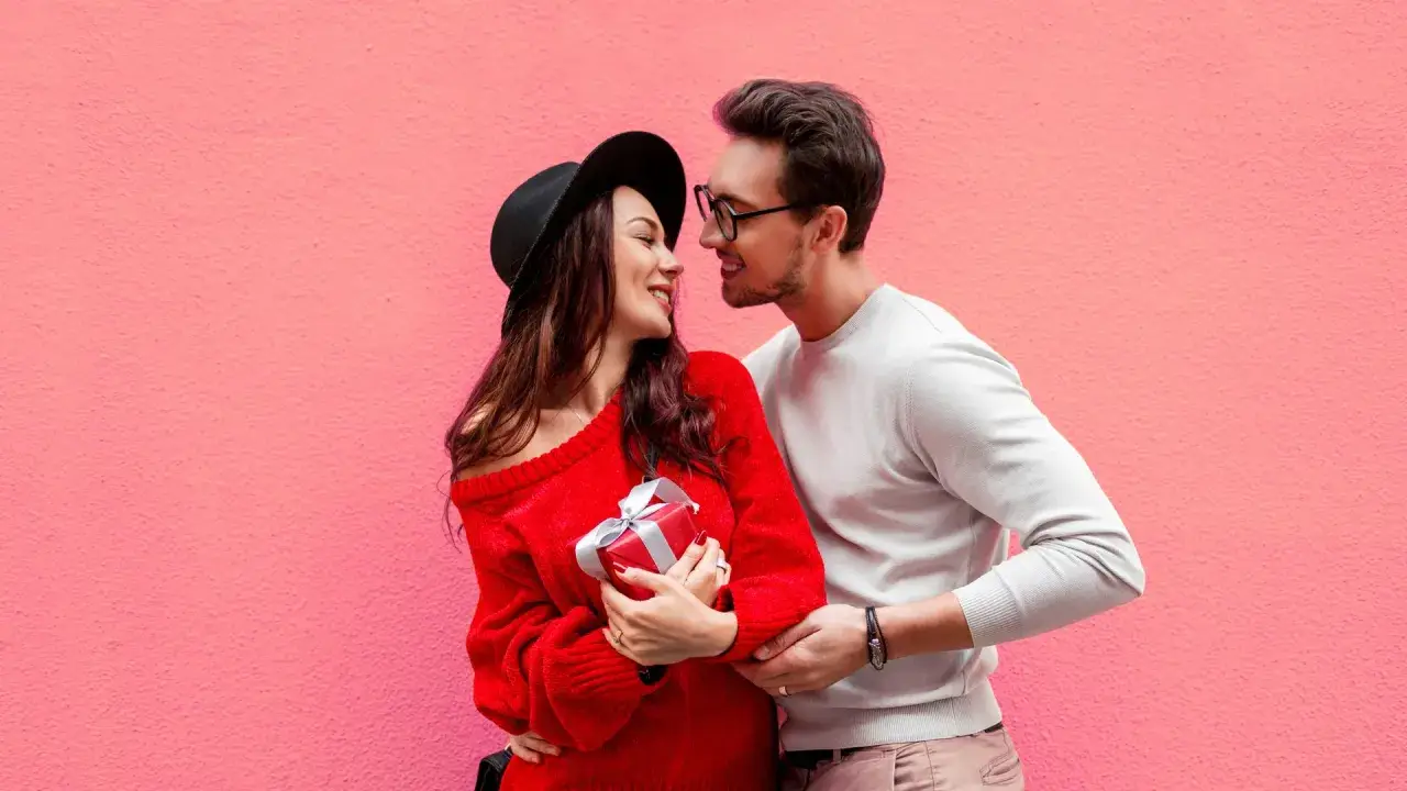 Para zakochanych na tle różowej ściany. Ona trzyma prezent, on obejmuje ją ramieniem. Mówi mu "I love you" po angielsku.