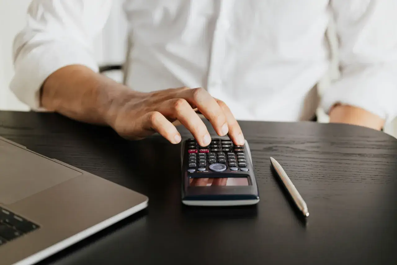 Dłoń mężczyzny nad kalkulatorem, obok laptop i długopis. Czy studia podyplomowe dają status studenta?