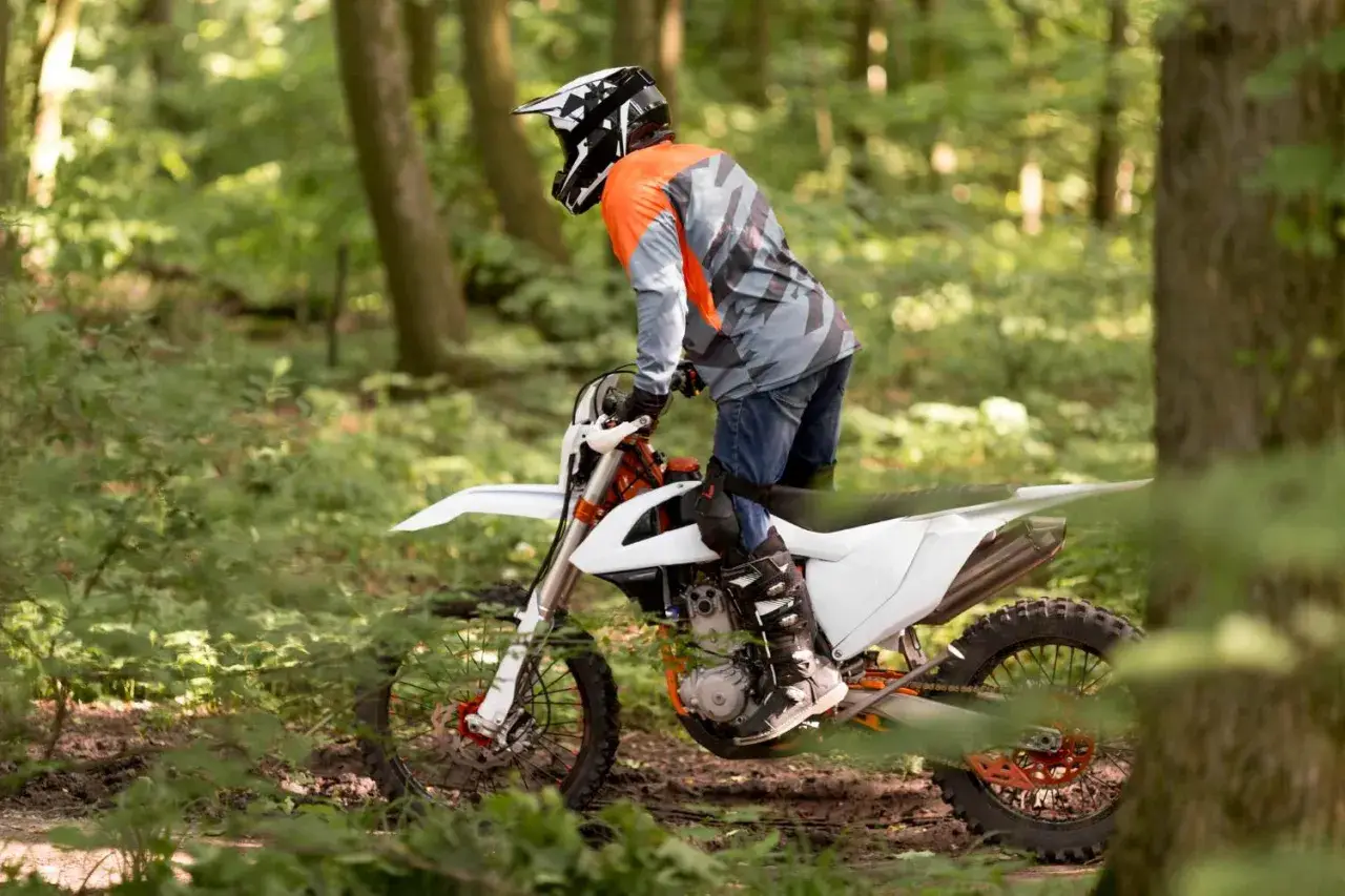 Motocyklista w kasku i pomarańczowej bluzie jedzie na białym crossie przez leśną ścieżkę.