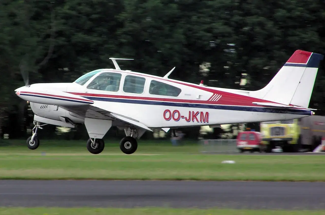 Eine weiße Beechcraft Bonanza mit roten und blauen Streifen hebt vom Gras ab.