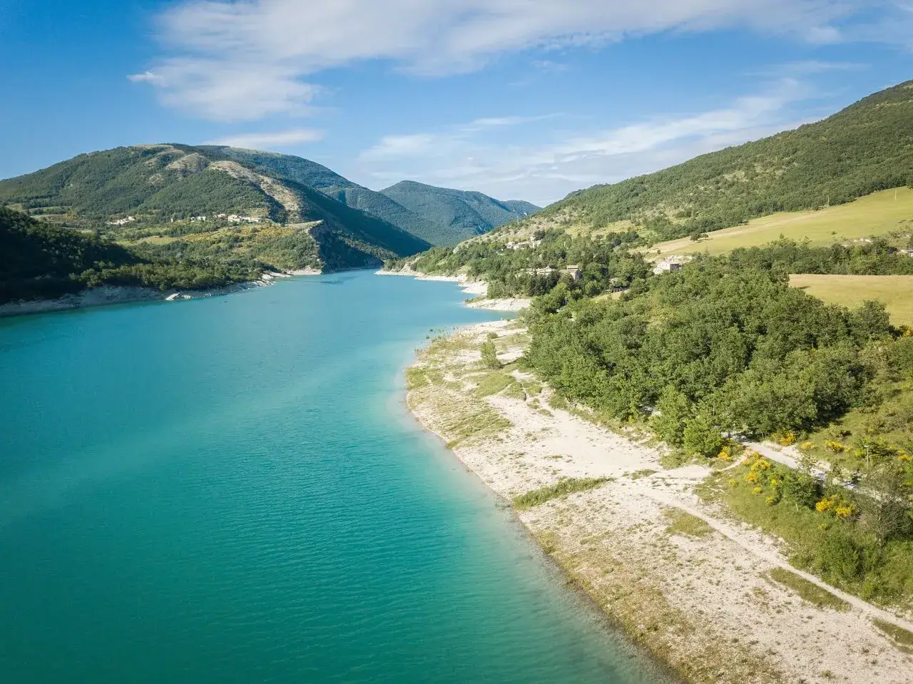 Splendido **lago di Fiastra** con acque turchesi, circondato da colline verdi e case sparse. Un paesaggio sereno e incantevole.