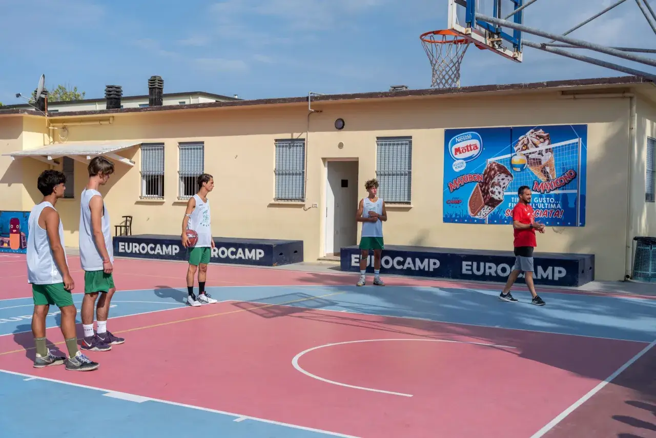 Ragazzi in tenuta sportiva giocano a basket in un camp estivo a Cesenatico, con un cartellone pubblicitario di gelati Motta sullo sfondo.