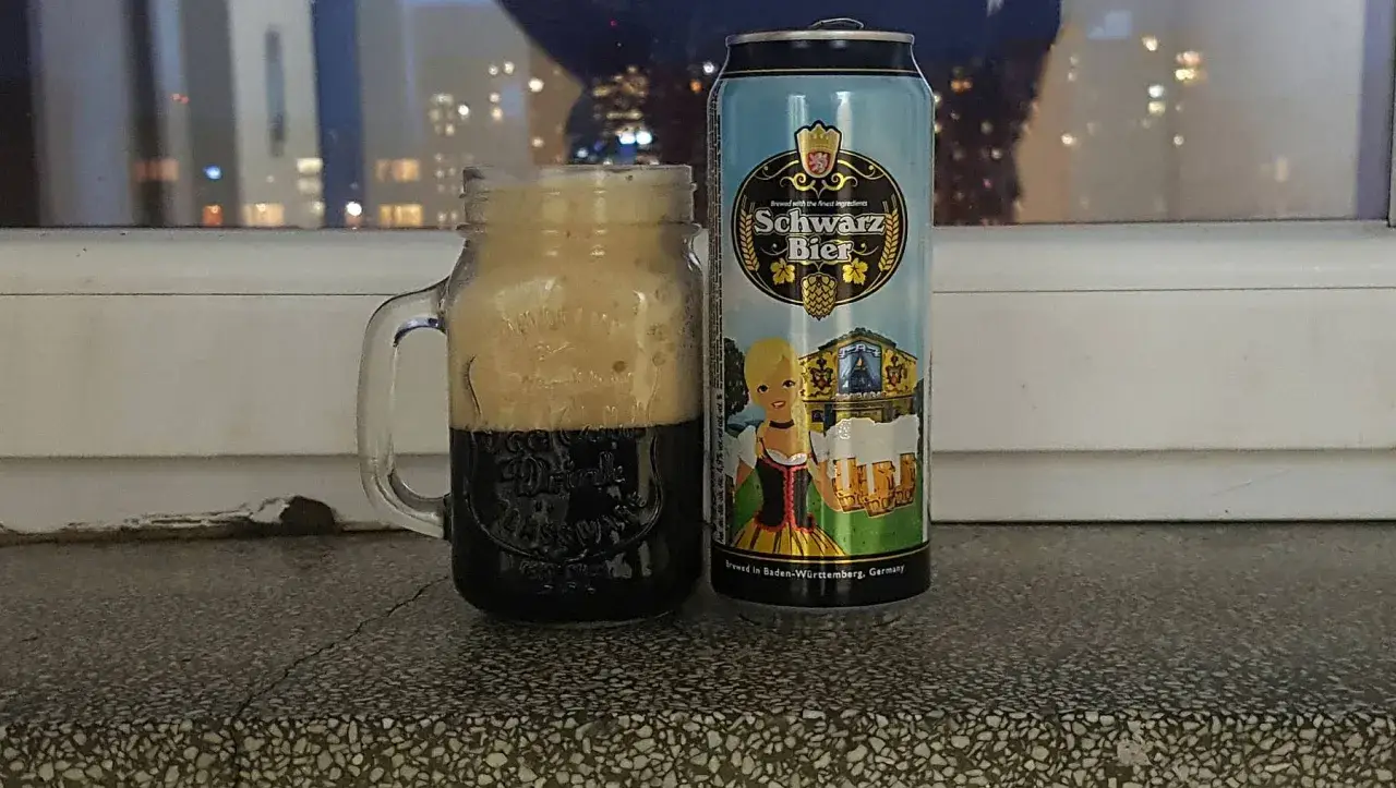 Perlenbacher Schwarz Bier w kuflu, obok puszka piwa. Wieczorny widok na miasto w tle.