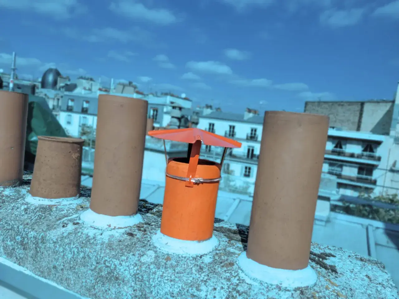 Sur un toit parisien, un chapeau chinois orange protège une cheminée, entouré d'autres conduits sous un ciel bleu.