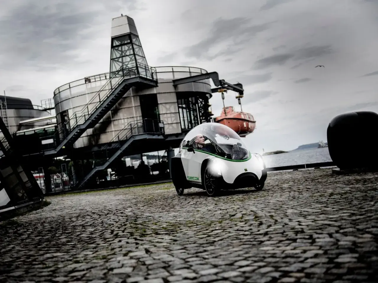 Ein Podbike Frikar fährt auf Kopfsteinpflaster vor einem modernen Gebäude am Wasser.