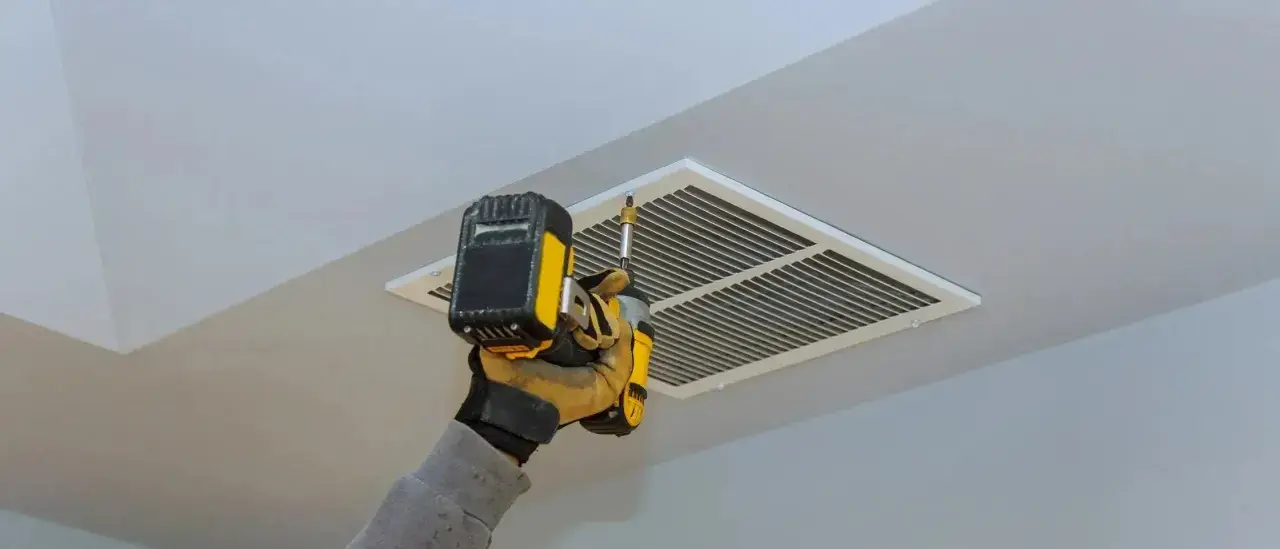Main tenant utilise une visseuse pour l'entretien de la VMC, fixant la grille d'aération au plafond.