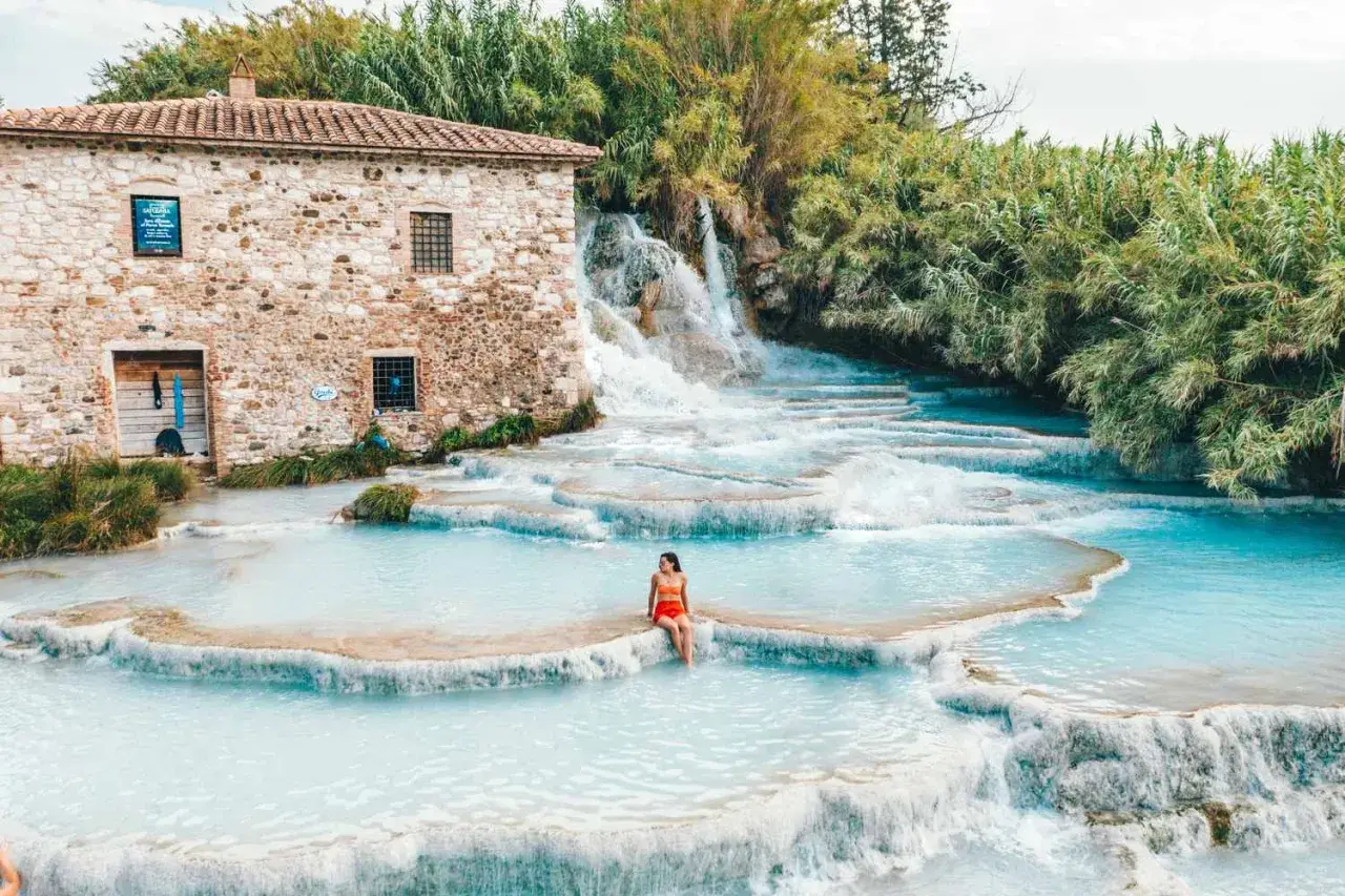 Cascate naturali con acqua turchese, una donna seduta su una roccia. Un antico mulino in pietra sullo sfondo. Tra le terme più belle d'Italia.