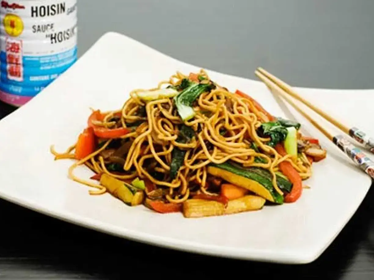 En tallrik med nudlar och grönsaker, perfekt för ett snabbt wok recept. Hoisinsåsburk syns i bakgrunden.