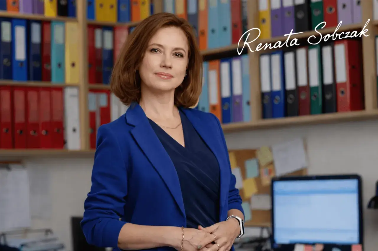Kobieta w niebieskiej marynarce stoi przed regałami z segregatorami.